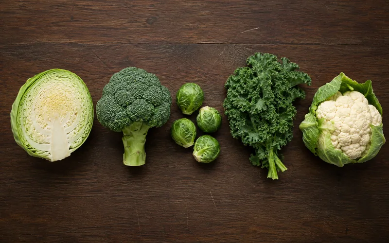 Five common brassica vegetables arranged on a dark wood surface including cabbage broccoli Brussels sprouts kale and cauliflower