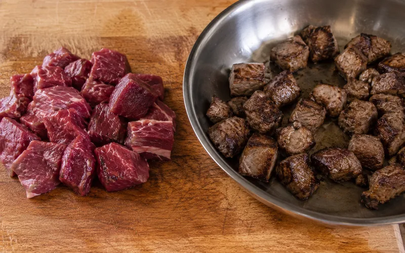 Cooked goat meat in a pan next to fresh raw goat meat on a cutting board for comparison