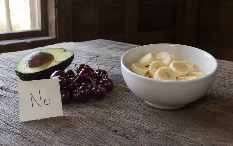 A wooden barn table showing an avocado and cherries marked off-limits beside a bowl of goat-safe banana pieces