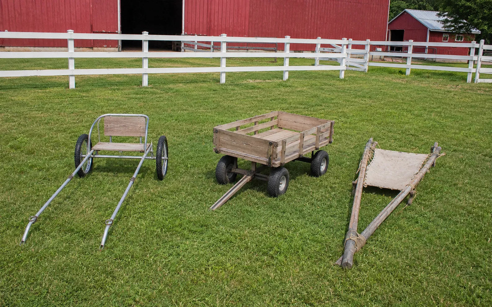 Three goat pulling vehicles side by side on a farm: a lightweight two-wheeled cart, a four-wheeled wooden wagon, and a simple travois