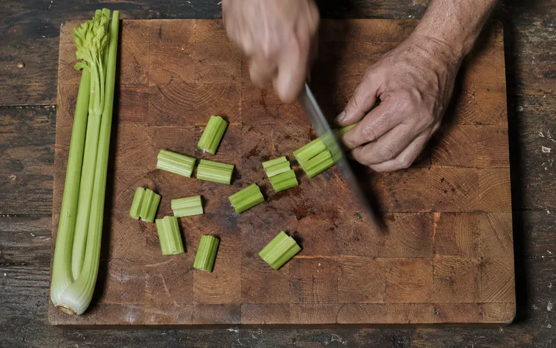 A person's hands chopping green celery stalks into short pieces on a wooden board