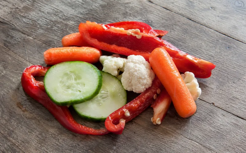 A colorful mix of goat-safe treats including red bell pepper strips, baby carrots, and cucumber rounds arranged on a rustic wooden table