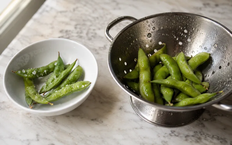 Frozen salted edamame pods in a bowl beside a colander of rinsed plain pods